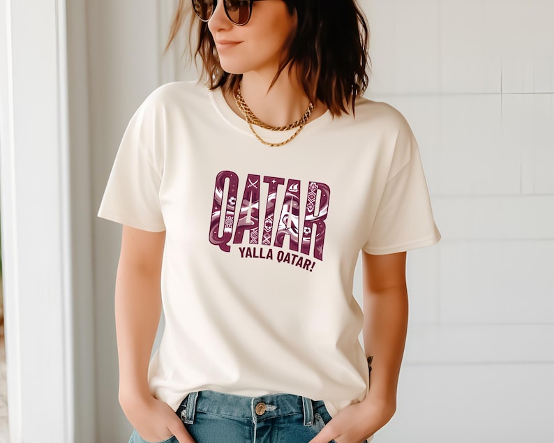 A woman wearing a white t-shirt with the text "QATAR YALLA QATAR!" printed on it.