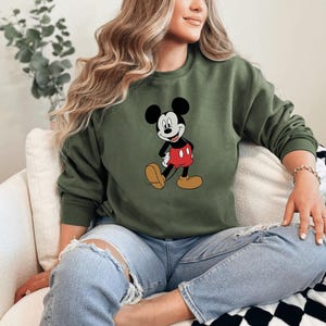 A woman wearing a green sweatshirt with a Mickey Mouse graphic is sitting on a couch.
