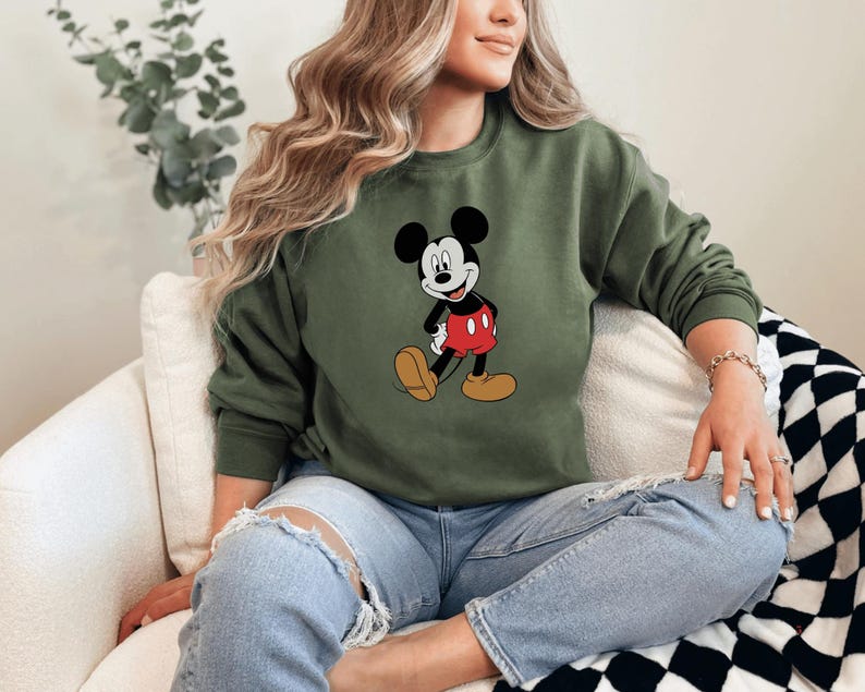A woman wearing a green sweatshirt with a Mickey Mouse graphic is sitting on a couch.