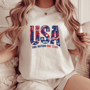 A woman wearing a white t-shirt with the text "USA" and "ONE NATION ONE TEAM" printed on it.