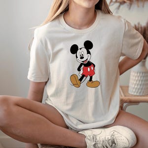 A woman wearing a white t-shirt with a Mickey Mouse graphic and white sneakers is sitting on a chair.