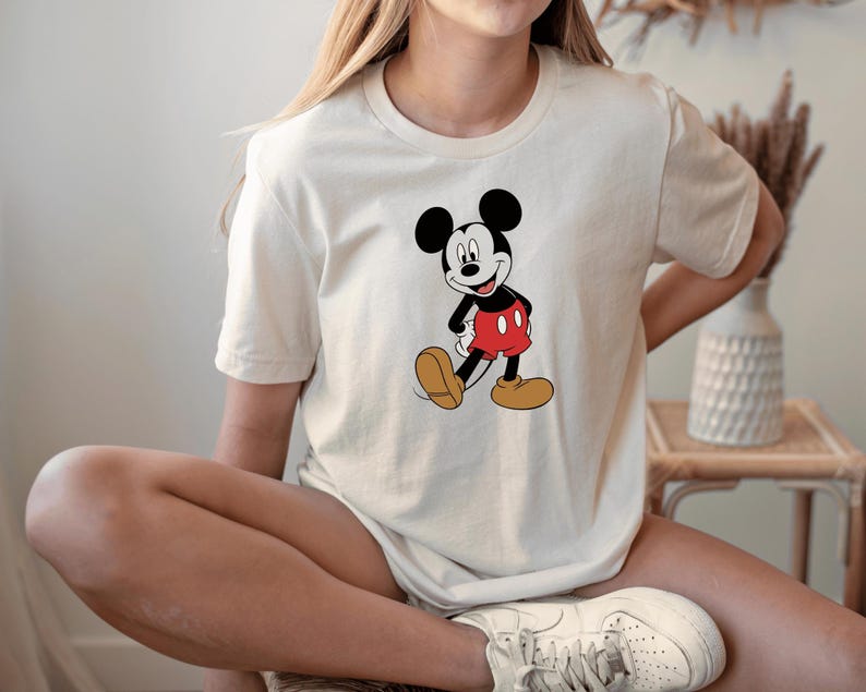 A woman wearing a white t-shirt with a Mickey Mouse graphic and white sneakers is sitting on a chair.