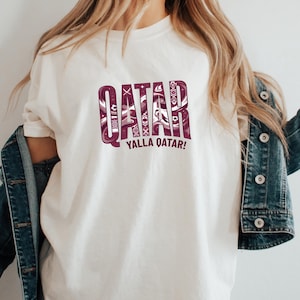 A person wearing a white t-shirt with the text "QATAR YALLA QATAR!" printed on it, along with a denim jacket.
