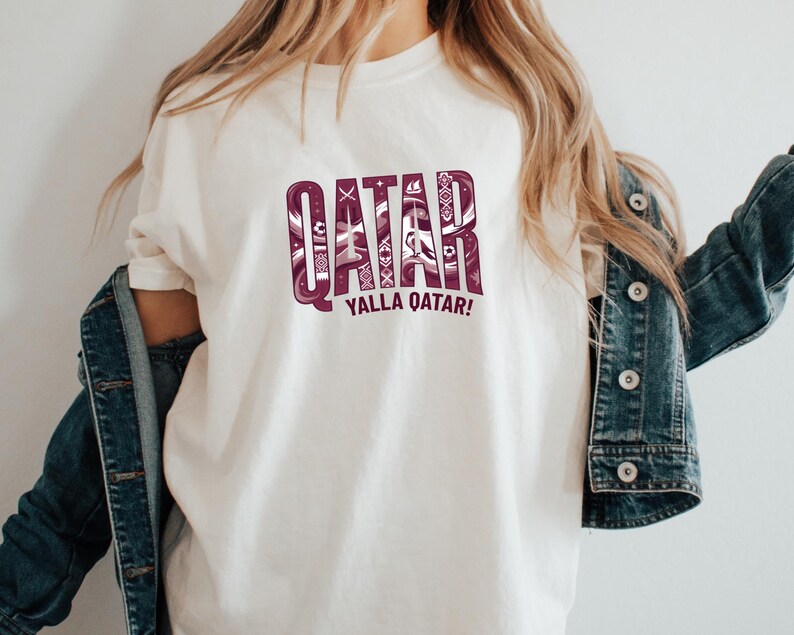 A person wearing a white t-shirt with the text "QATAR YALLA QATAR!" printed on it, along with a denim jacket.