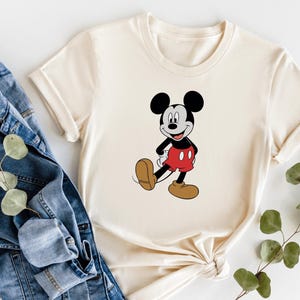 A white t-shirt with a cartoon image of Mickey Mouse, a classic Disney character, is displayed alongside a pair of blue jeans and some greenery.