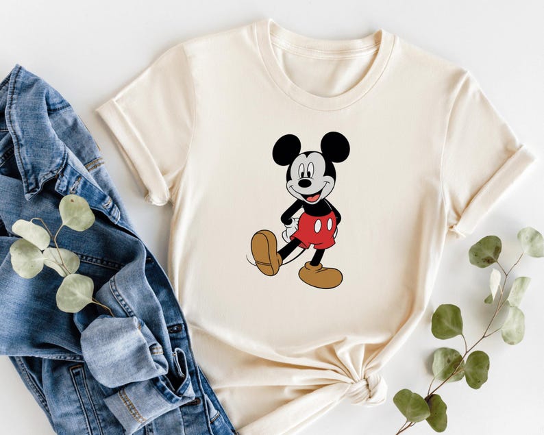 A white t-shirt with a cartoon image of Mickey Mouse, a classic Disney character, is displayed alongside a pair of blue jeans and some greenery.