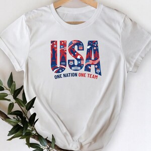 A white t-shirt with the text "USA" and "ONE NATION ONE TEAM" printed on it, along with various patriotic symbols and designs.