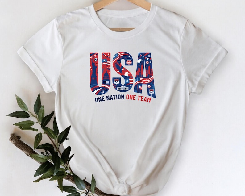 A white t-shirt with the text "USA" and "ONE NATION ONE TEAM" printed on it, along with various patriotic symbols and designs.