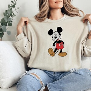 A woman is sitting on a couch, wearing a white sweater with a Mickey Mouse graphic, and has her hands on her shoulders.