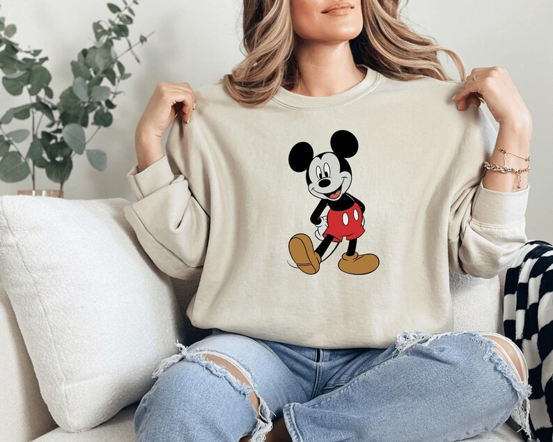 A woman is sitting on a couch, wearing a white sweater with a Mickey Mouse graphic, and has her hands on her shoulders.