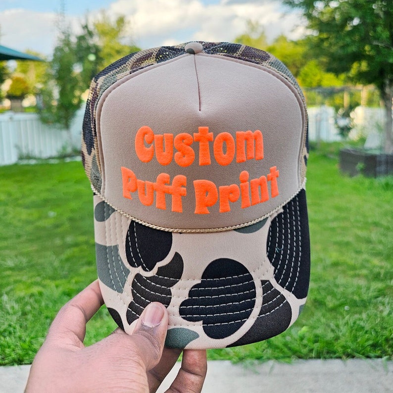 Puede incluir: Gorra de camionero color beige con un estampado de camuflaje en la parte delantera y una parte trasera de malla. La gorra presenta las palabras "Custom Puff Print" en naranja brillante.