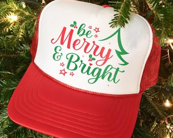 May include: A red and white trucker hat with the words "be Merry & Bright" in red and green script. The hat features a red brim and mesh back, with a Christmas tree graphic.