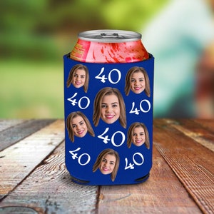 May include: A blue can cooler featuring a repeating pattern of a woman's face and the number "40" in white. The cooler is holding a red and silver can. The background is blurred, with a wooden surface in the foreground.