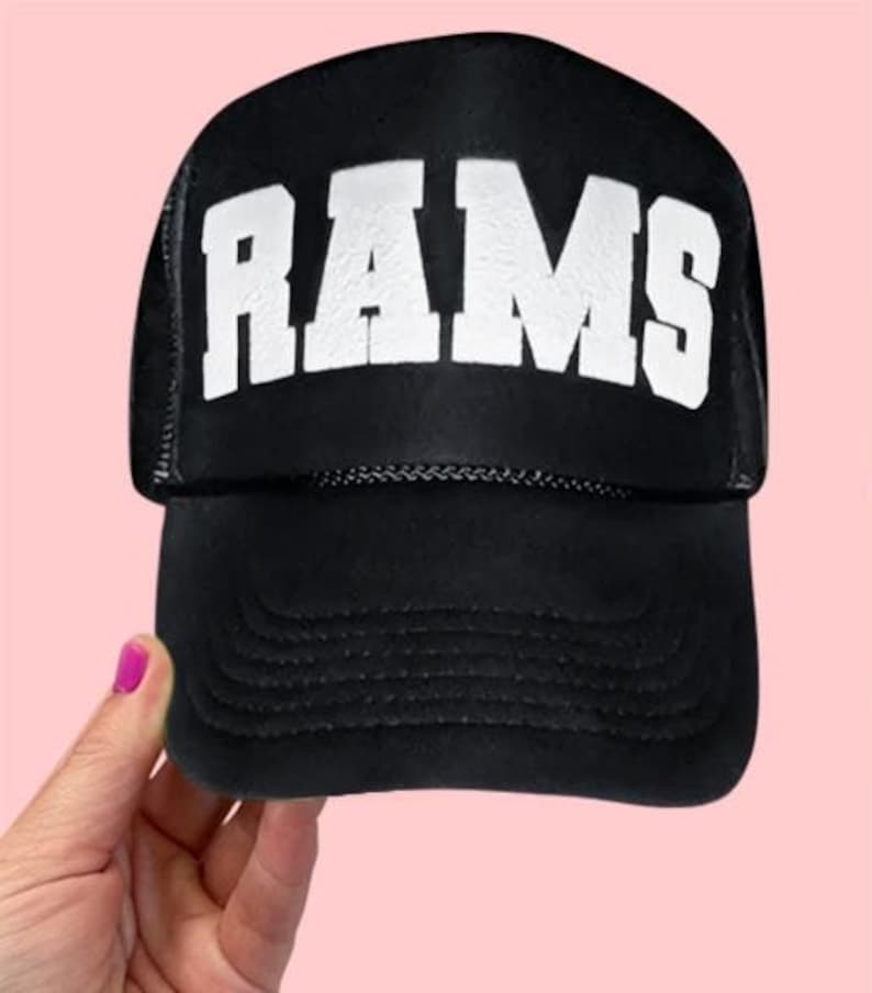 May include: Black trucker hat with the word "RAMS" in large, white, block letters across the front. The hat has a curved bill and a mesh back. The hat is being held by a person with pink nail polish.