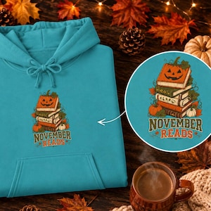 Może przedstawiać: Turkusowa bluza z kapturem z grafiką przedstawiającą stos książek zwieńczony dynią na Halloween, otoczony jesiennymi liśćmi i małymi dyniami. Poniżej stosu znajduje się napis "NOVEMBER READS" wykonany ozdobną, stylizowaną czcionką.