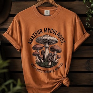 May include: A burnt orange short-sleeve t-shirt featuring a graphic of various mushrooms and the text "AMATEUR MYCOLOGIST" arched above and "WITH QUESTIONABLE MORELS" arched below the illustration in a dark brown, bold, sans-serif font.