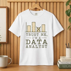 May include: A white short-sleeve t-shirt featuring a graphic of four bar charts, with the third bar marked by an X. Below the graphic, the text reads: TRUST ME, I'M A DATA ANALYST. The design is printed in a distressed, dark olive green color.
