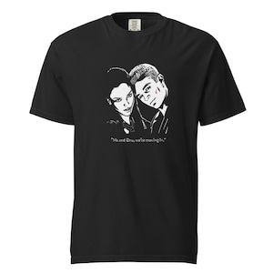 May include: A black short-sleeved t-shirt featuring a white graphic illustration of two people with the text "Me and Dru, we're moving in." printed below the image. The shirt has a crew neckline and a standard fit, made of solid black fabric.
