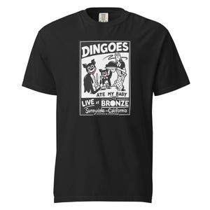 May include: A black short-sleeved t-shirt featuring a white graphic print of two cartoon dingoes in suits at a table with a server. The text reads: DINGOES ATE MY BABY, LIVE at THE BRONZE, Sunnydale, California. The shirt has a crew neckline.