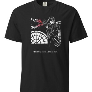 May include: A black short-sleeved t-shirt featuring a white graphic illustration of a person aiming a weapon with a red muzzle flash. The design includes a circular window background and the printed text, "That was then... this is now."