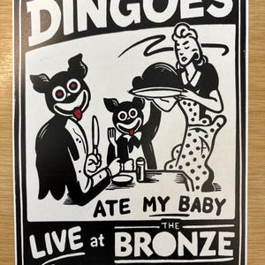 May include: A black and white graphic print featuring two cartoon dingoes in suits sitting at a table with a person in a polka dot dress. The text reads DINGOES ATE MY BABY LIVE at THE BRONZE Sunnydale-California in bold black lettering.