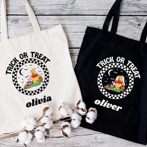May include: Two white canvas tote bags with black handles. The bags have a black and white checkered circle with the text "Trick or Treat" and a cartoon image of Winnie the Pooh and Piglet. One bag has the name "Olivia" printed below the circle and the other bag has the name "Oliver" printed below the circle.