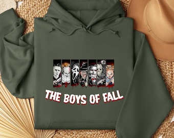May include: An olive green hooded sweatshirt featuring a graphic print of seven horror movie characters arranged in vertical panels. Below the characters, the text THE BOYS OF FALL is printed in a white, blood-dripping font style on the chest.