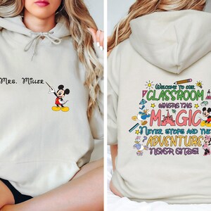 May include: A light beige hoodie with a Mickey Mouse illustration on the front and text that says "MRS. MILLER". The back of the hoodie has a colorful graphic with Mickey Mouse, Donald Duck, and Minnie Mouse characters and text that says "Welcome to our classroom where the magic never stops and the adventure never ends!"