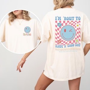 May include: Cream-colored t-shirt with a large graphic on the back that says "I'm 'bout to have a good day." The graphic includes a winking smiley face, flowers, and a checkered pattern in pink and blue.