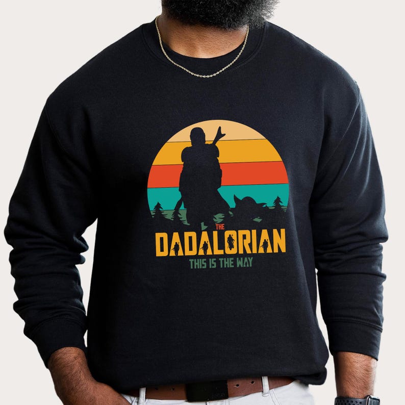 Puede incluir: Sudadera negra con un gr&aacute;fico de una figura silueta y una figura infantil frente a una puesta de sol. El texto dice "THE DADALORIAN THIS IS THE WAY" en amarillo y verde. La sudadera es de cuello redondo.