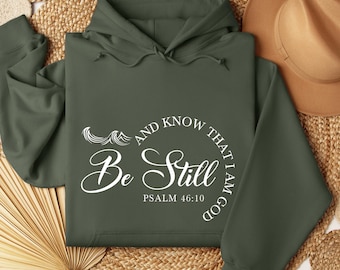 May include: A dark green hooded sweatshirt with the text "Be Still and Know That I Am God" in white lettering. The text is in a circle with a wave design above it. The text "Psalm 46:10" is below the circle.