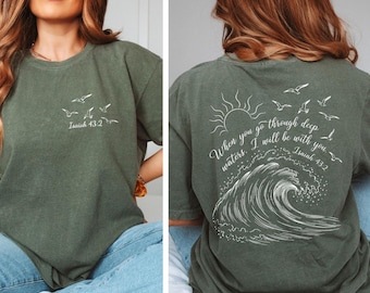 May include: Olive green t-shirt with white graphic text and illustrations. The front features flying birds and the text "Isaiah 43:2". The back displays a sun, wave, birds, and the text "When you go through deep waters, I will be with you. Isaiah 43:2".