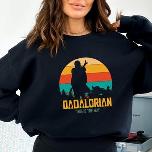 Puede incluir: Sudadera negra con un dise&ntilde;o gr&aacute;fico. El dise&ntilde;o presenta la silueta de una persona y una peque&ntilde;a criatura sobre un fondo de puesta de sol. El texto dice "The Dadalorian, This is the Way" en amarillo.