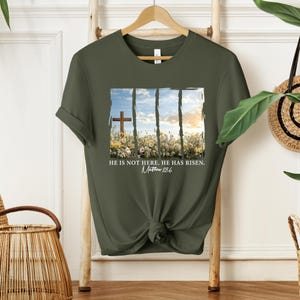 May include: A green t-shirt with a white graphic of a cross in a field of flowers. The graphic has a white border and the text "He is not here. He has risen. Matthew 28:6".
