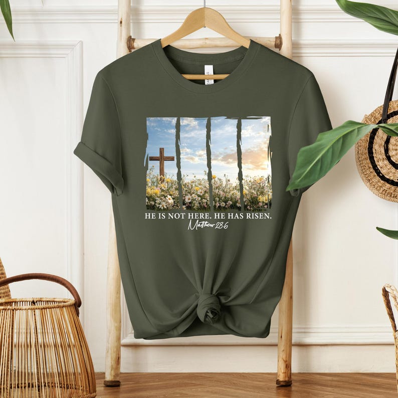 May include: A green t-shirt with a white graphic of a cross in a field of flowers. The graphic has a white border and the text "He is not here. He has risen. Matthew 28:6".