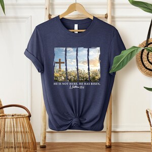 May include: Navy blue t-shirt with a graphic of a cross in a field of flowers. The text "He is not here. He has risen. Matthew 28:6" is printed below the graphic.