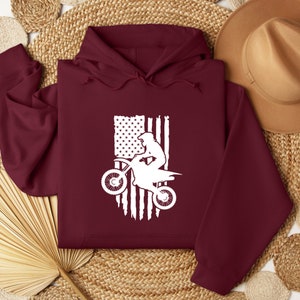 May include: Burgundy hooded sweatshirt with a white silhouette of a person riding a dirt bike in front of a distressed American flag.
