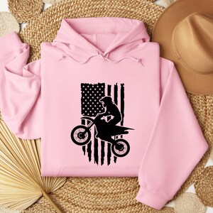 May include: Pink hooded sweatshirt with a black silhouette of a person riding a dirt bike in front of an American flag.
