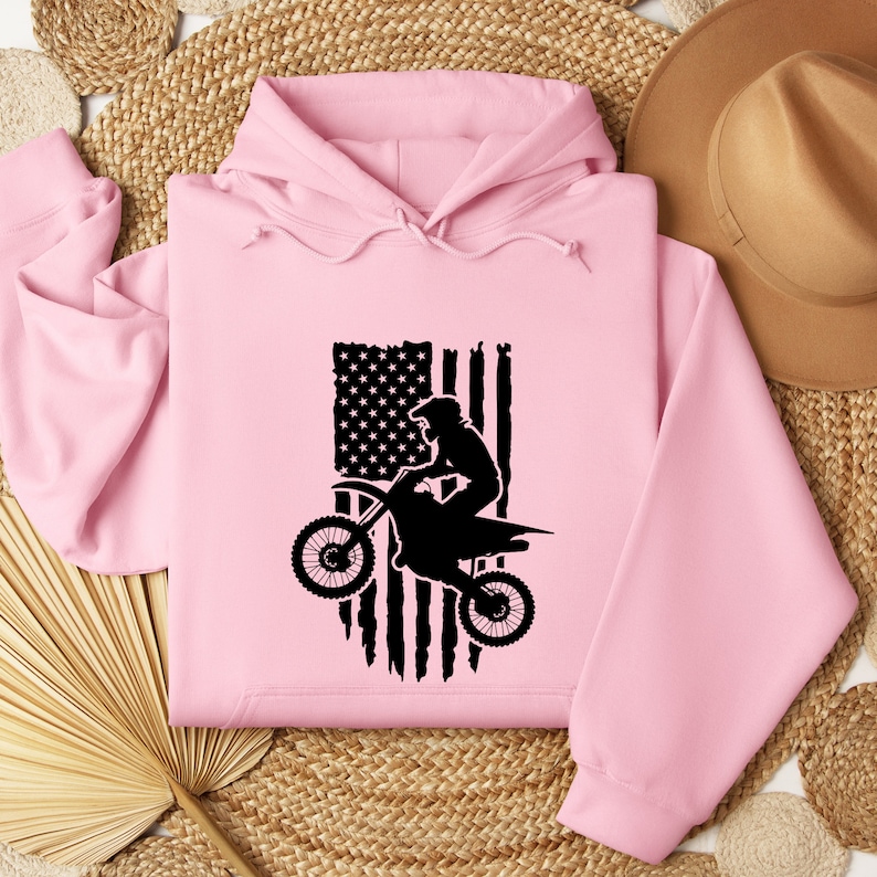 May include: Pink hooded sweatshirt with a black silhouette of a person riding a dirt bike in front of an American flag.
