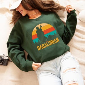 Puede incluir: Sudadera verde oscuro con un dise&ntilde;o retro de puesta de sol. El dise&ntilde;o presenta la silueta de una figura y el texto "The Dadalorian" en dorado, con "This is the Way" debajo. La sudadera es de color verde oscuro.