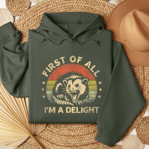 May include: Dark green hoodie with a graphic of an opossum inside a circle with the text "FIRST OF ALL I'M A DELIGHT". The opossum graphic is in shades of tan, orange, and yellow. The hoodie has a drawstring hood and long sleeves.