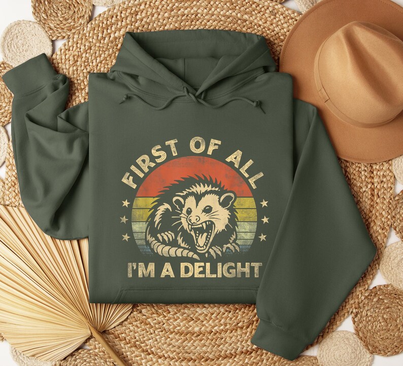 May include: Dark green hoodie with a graphic of an opossum inside a circle with the text "FIRST OF ALL I'M A DELIGHT". The opossum graphic is in shades of tan, orange, and yellow. The hoodie has a drawstring hood and long sleeves.