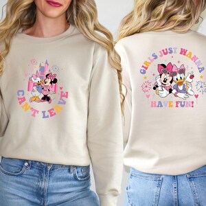 Puede incluir: Una sudadera de color crema con un gr&aacute;fico de Mickey Mouse y Daisy Duck frente a un castillo rosa. El texto "Can't Leave" est&aacute; escrito en un c&iacute;rculo alrededor del gr&aacute;fico. La parte posterior de la sudadera tiene un gr&aacute;fico de Mickey Mouse y Daisy Duck con el texto "Girls Just Wanna Have Fun!" en un c&iacute;rculo alrededor del gr&aacute;fico.