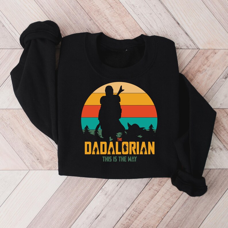 Puede incluir: Sudadera negra con un gr&aacute;fico de una silueta de figura y ni&ntilde;o contra un atardecer. El texto dice "The Dadalorian" y "This is the Way" en amarillo y blanco.