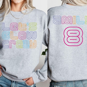 May include: A light gray sweatshirt with the text "LET'S GLOW CRAZY" in a colorful, retro font on the front. The back of the sweatshirt features the name "EMILY" and the number "8" in a similar style.
