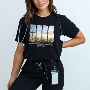 May include: Black t-shirt with a graphic of a cross in a field of flowers. The text "He is not here. He has risen. Matthew 28:6" is printed below the graphic.