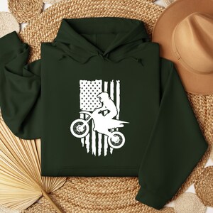 May include: A dark green hoodie with a white silhouette of a person riding a dirt bike in front of an American flag.