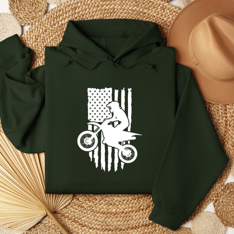 May include: A dark green hoodie with a white silhouette of a person riding a dirt bike in front of an American flag.