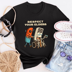 May include: Black t-shirt with the text "RESPECT YOUR ELDERS" and a cartoon graphic of a video game controller pushing a retro game controller in a wheelchair. White sneakers, denim shorts, and accessories are also in the image.