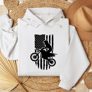 May include: White hooded sweatshirt with a black silhouette of a motocross rider jumping over a distressed American flag.